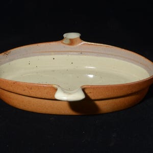 May include: A handcrafted ceramic dish, featuring a warm brown exterior and a cream-coloured interior. The dish has a distinctive shape with a small spout and a slightly raised rim. The design is simple and elegant, suitable for serving or display, with a diameter of approximately 20 cm.
