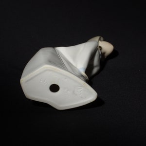 May include: White porcelain figurine of a woman in a white dress with blue accents. The figurine is sitting down with her head bowed. The bottom of the figurine has a mark that reads "Lladro" and "Spain".