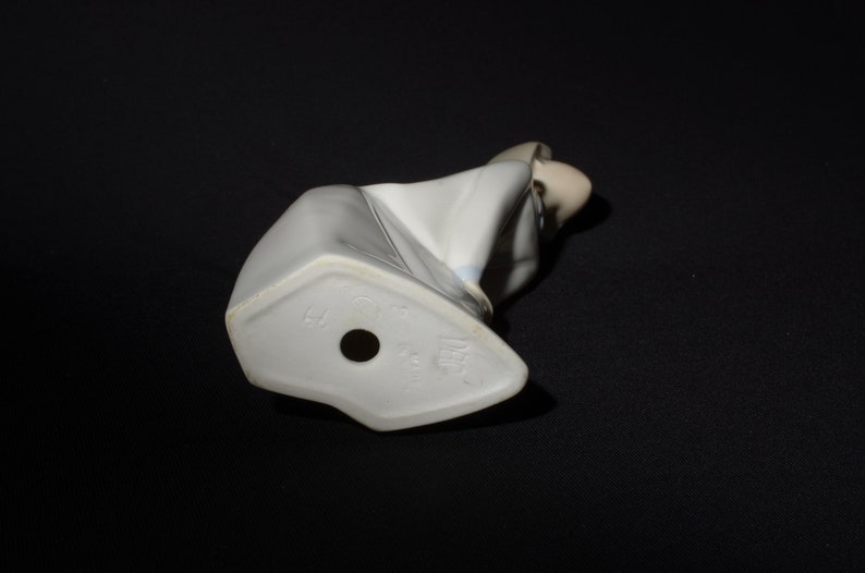 May include: White porcelain figurine of a woman in a white dress with blue accents. The figurine is sitting down with her head bowed. The bottom of the figurine has a mark that reads "Lladro" and "Spain".