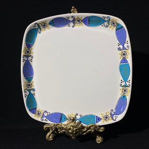 Può includere: Un piatto quadrato in ceramica bianca con un bordo di disegni stilizzati di pesci blu e turchese. I pesci sono intervallati da elementi floreali gialli e neri. Il piatto è esposto su un supporto decorativo in ottone.