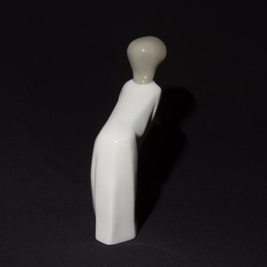 May include: White porcelain figurine of a person in a long dress with a gray head. The figurine is standing on a black surface.