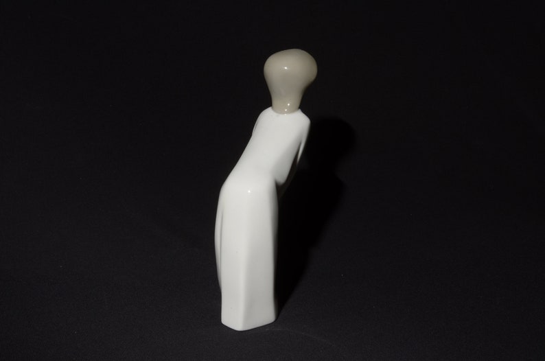 May include: White porcelain figurine of a person in a long dress with a gray head. The figurine is standing on a black surface.