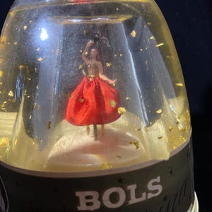 May include: A clear glass bottle with a gold liquid and gold flakes. Inside, a ballerina figurine in a red dress is poised on a white base. The bottle has a black label with the word "BOLS".