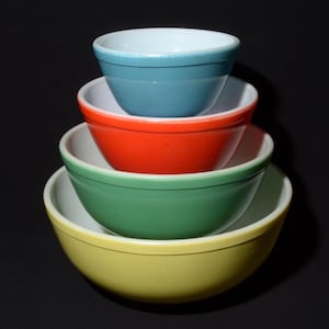 May include: A set of four nested mixing bowls with a smooth finish and white interiors. The bowls are stacked from smallest to largest, featuring a teal blue top bowl, a bright orange second bowl, a sage green third bowl, and a yellow base bowl.