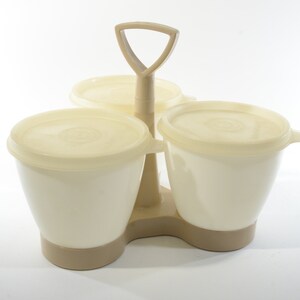 May include: A set of three off-white plastic containers with lids, attached to a beige plastic handle. The containers are designed for storing food.