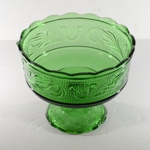 May include: A green glass pedestal bowl with a scalloped rim and intricate floral designs. The bowl has a footed base.