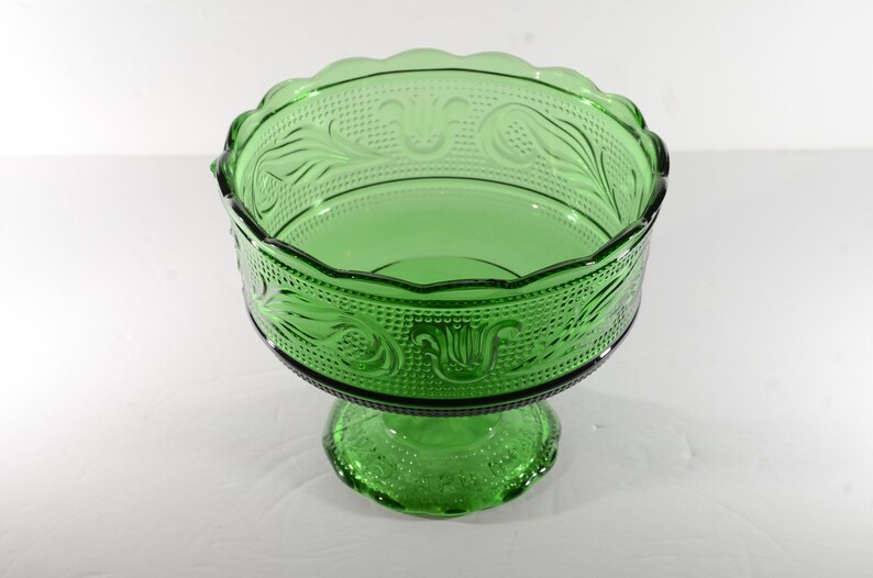 May include: A green glass pedestal bowl with a scalloped rim and intricate floral designs. The bowl has a footed base.