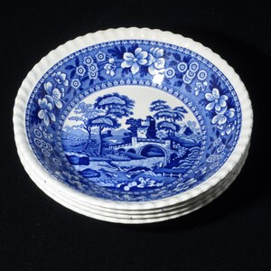 Può includere: Una pila di ciotole in ceramica blu e bianca. Ogni ciotola presenta una scena paesaggistica dettagliata con un ponte e alberi. I bordi sono decorati con motivi floreali e un bordo smerlato.