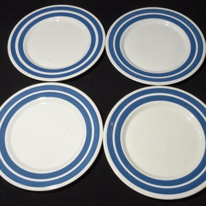 May include: Four white ceramic plates with a blue double-stripe design around the rim. The plates are arranged in a square formation, showcasing a classic and clean aesthetic. Suitable for everyday dining or special occasions.