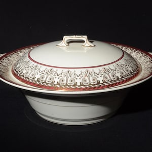 Może przedstawiać: Biała ceramiczna miska do serwowania z pokrywką. Miska ma czerwoną i złotą obwódkę z ozdobnym wzorem. Pokrywka ma złoty uchwyt.