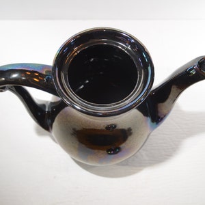 May include: A black iridescent ceramic teapot with a rounded body and a curved spout. The teapot has a shiny, metallic finish.