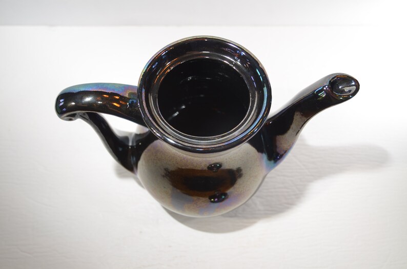 May include: A black iridescent ceramic teapot with a rounded body and a curved spout. The teapot has a shiny, metallic finish.
