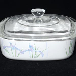 Pode incluir: Travessa branca com tampa de vidro transparente. A travessa apresenta um desenho floral de íris azuis e roxas com caules verdes. A travessa tem duas pegas brancas em cada lado.