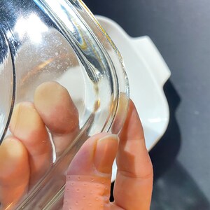 K&ouml;nnte beinhalten: Nahaufnahme eines durchsichtigen Glasdeckels f&uuml;r eine Auflaufform. Der Deckel hat einen leicht abgerundeten Rand und wird von einer Hand gehalten.