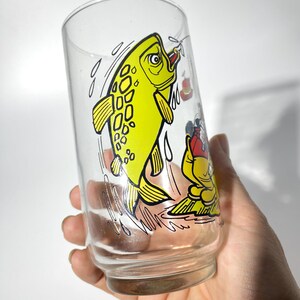 K&ouml;nnte beinhalten: Klares Glas mit einer Cartoon-Illustration eines gelben Fisches, der aus dem Wasser springt. Der Fisch hat schwarze Umrisse und Details. Eine kleinere Cartoon-Figur ist ebenfalls zu sehen. Das Glas wird von einer Hand gehalten.