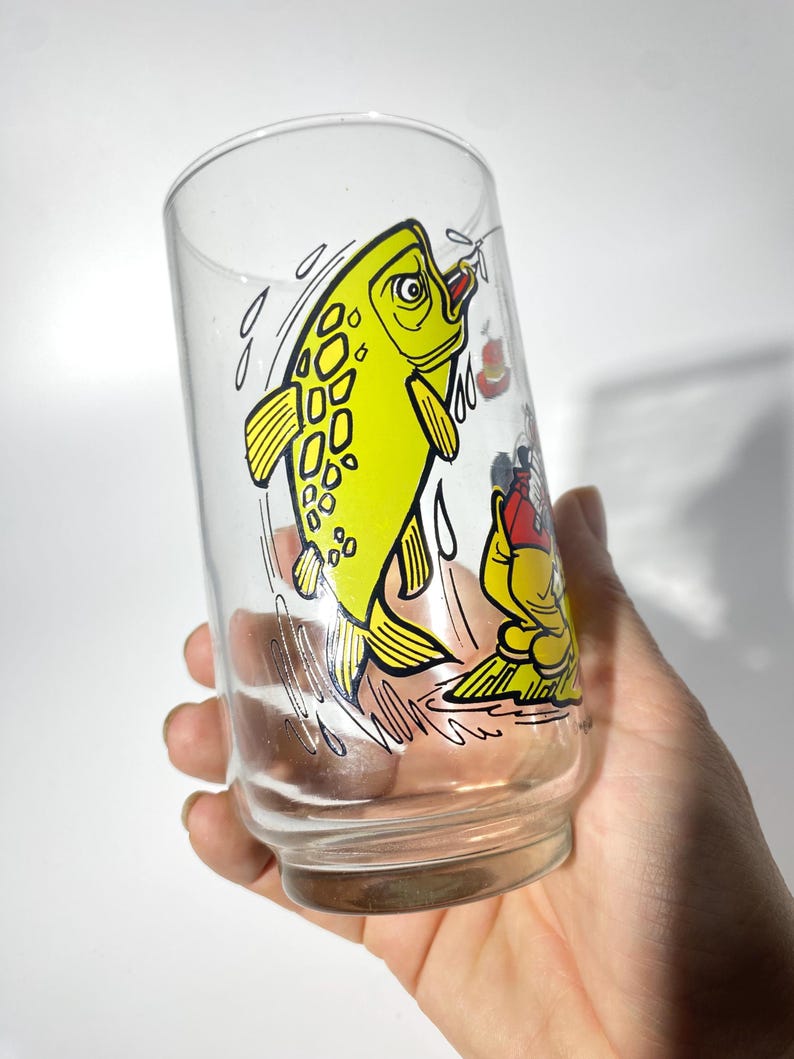 K&ouml;nnte beinhalten: Klares Glas mit einer Cartoon-Illustration eines gelben Fisches, der aus dem Wasser springt. Der Fisch hat schwarze Umrisse und Details. Eine kleinere Cartoon-Figur ist ebenfalls zu sehen. Das Glas wird von einer Hand gehalten.