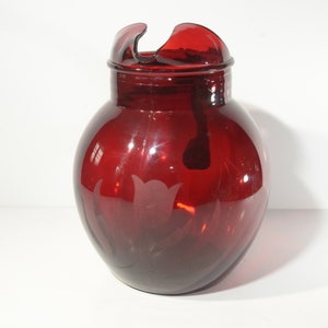 Puede incluir: Una jarra de vidrio roja con una boca ancha y un asa curva. La jarra tiene un diseño esmerilado de tulipanes en la parte delantera.