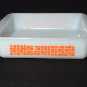 May include: A white rectangular Pyrex baking dish with rounded corners. It has an orange geometric pattern along one side, made up of squares and circles. The dish is empty and rests on a black surface.