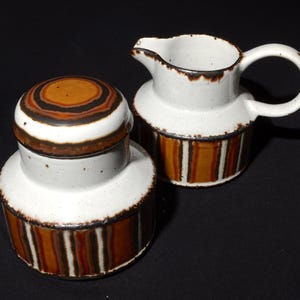 Könnte beinhalten: Ein weißes Zucker- und Milchkännchen-Set aus Keramik mit einem braunen, orangefarbenen und schwarzen Streifenmuster. Der Deckel der Zuckerdose hat ein braunes, orangefarbenes und schwarzes konzentrisches Kreismuster.
