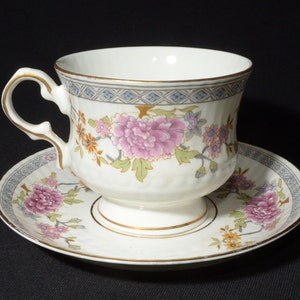 Puede incluir: Una taza de té y un platillo blancos con un patrón floral. La taza tiene un borde dorado y el platillo tiene una banda dorada alrededor del borde. Las flores son rosas, amarillas y azules.