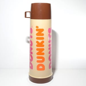 May include: A vintage Dunkin' Donuts thermos. The thermos is beige with brown accents on the lid and base. The Dunkin' Donuts logo is printed vertically in orange and pink. The thermos has a handle on the lid.