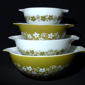 May include: Four vintage Pyrex mixing bowls stacked on top of each other. The bowls are white and olive green with a floral design. Each bowl has a handle on either side.