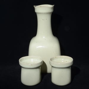 May include: A cream-colored ceramic sake set, including a tall, slender bottle and two small, cylindrical cups. The bottle has a rounded body and a flared top. The set is displayed against a dark background.