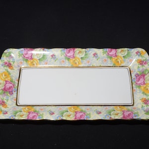 Peut inclure: Un plateau de service rectangulaire avec un bord festonné. Le plateau est décoré d'un motif floral dans des tons de rose, jaune et bleu sur fond blanc. Le plateau est bordé d'une garniture dorée.