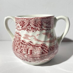 May include: A white ceramic creamer with a red floral and landscape design. The creamer has two handles and a rounded body.