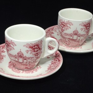 May include: Two white teacups and saucers with a red floral and architectural design. The cups have a handle and sit on matching saucers. The design includes buildings and floral patterns. The set is on a black background.