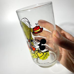 K&ouml;nnte beinhalten: Klares Glas mit einem Vintage-Mickey-Mouse-Design. Der Cartoon zeigt Mickey Mouse beim Angeln, mit einem rot-gelben Hut. Das Glas tr&auml;gt unten den Text "Walt Disney Productions".