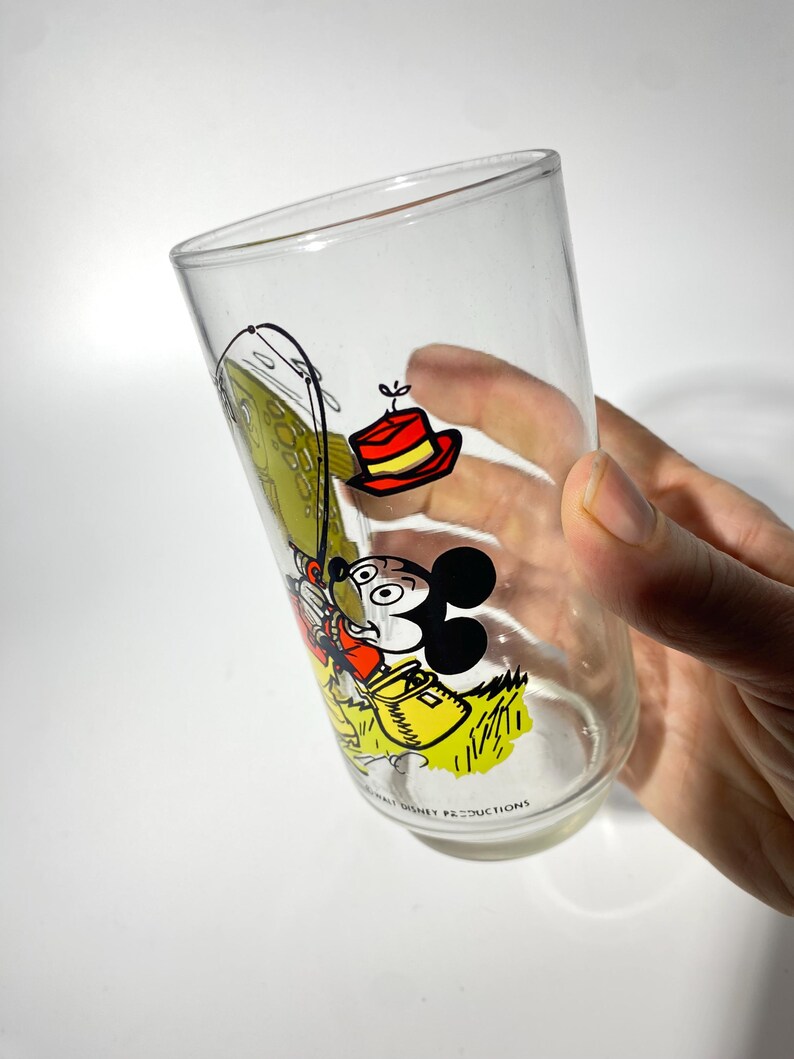 K&ouml;nnte beinhalten: Klares Glas mit einem Vintage-Mickey-Mouse-Design. Der Cartoon zeigt Mickey Mouse beim Angeln, mit einem rot-gelben Hut. Das Glas tr&auml;gt unten den Text "Walt Disney Productions".