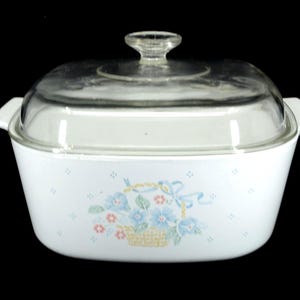 Op de afbeelding: Witte ovenschaal met een helder glazen deksel. De schaal heeft een bloemenontwerp met blauwe, roze en gele accenten. Het heeft twee handvatten en is geschikt om in te bakken en te serveren.