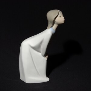 May include: A white porcelain figurine of a girl with brown hair, wearing a white dress with a blue collar, looking down and to the side.
