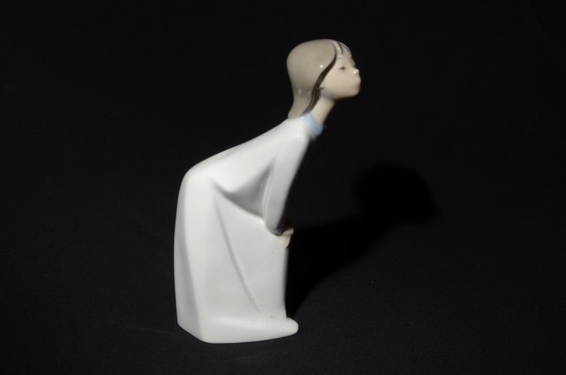 May include: A white porcelain figurine of a girl with brown hair, wearing a white dress with a blue collar, looking down and to the side.