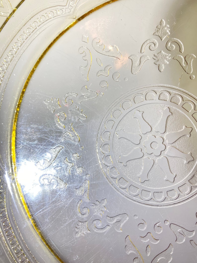 May include: A vintage glass plate with a yellow rim and a white floral design. The plate has a center design of a wheel with a flower in the middle.