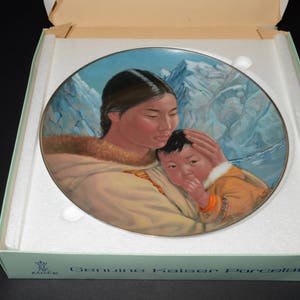 May include: Kaiser Porcelain decorative plate depicting a mother and child in traditional attire, set against a backdrop of snow-covered mountains. The plate is in its original box, with the words "Genuine Kaiser Porcelain" visible.