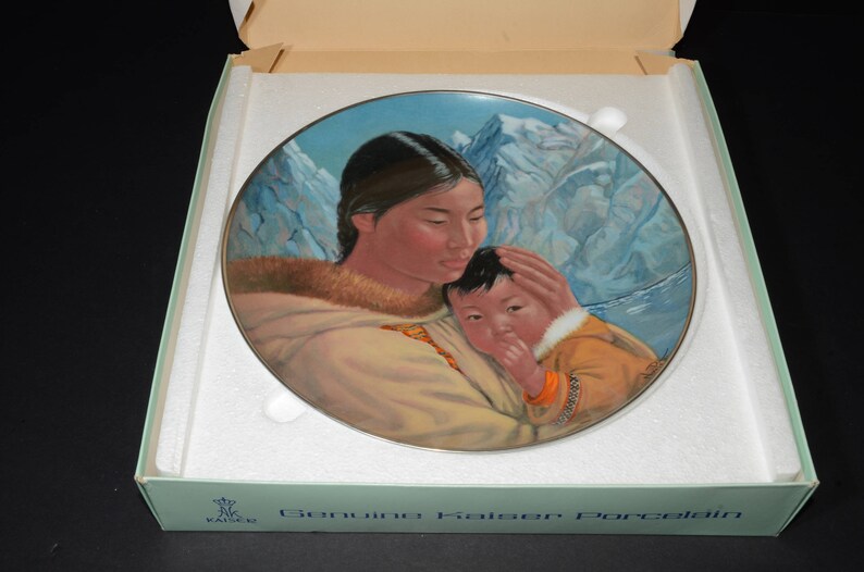 May include: Kaiser Porcelain decorative plate depicting a mother and child in traditional attire, set against a backdrop of snow-covered mountains. The plate is in its original box, with the words "Genuine Kaiser Porcelain" visible.
