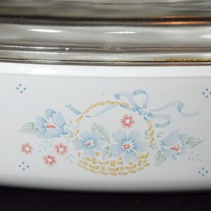 K&ouml;nnte beinhalten: Wei&szlig;e Keramik-Backform mit einem Blumendesign mit blauen Blumen, gr&uuml;nen Bl&auml;ttern und einem Korb mit einem gelben und braunen gewebten Muster. Die Form hat einen Glasdeckel.