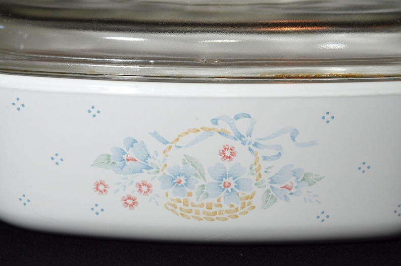 K&ouml;nnte beinhalten: Wei&szlig;e Keramik-Backform mit einem Blumendesign mit blauen Blumen, gr&uuml;nen Bl&auml;ttern und einem Korb mit einem gelben und braunen gewebten Muster. Die Form hat einen Glasdeckel.