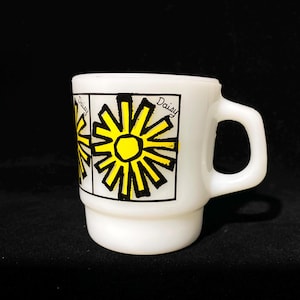 May include: White ceramic mug featuring a yellow and black daisy design. The mug has a handle and the word "Daisy" is written on the side. The mug is a vintage style and is set against a black background.