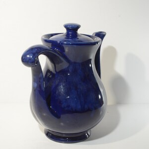 May include: A cobalt blue ceramic teapot with a unique, curved spout and handle. The teapot has a glossy finish and a slightly textured surface.