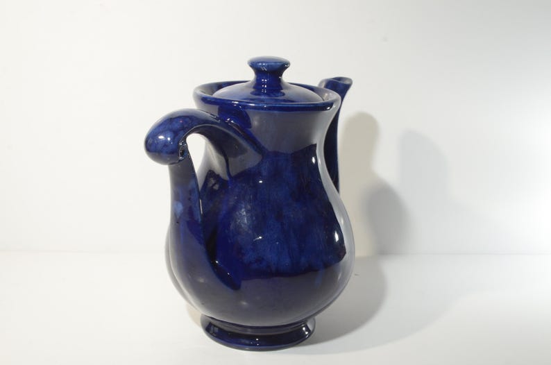 May include: A cobalt blue ceramic teapot with a unique, curved spout and handle. The teapot has a glossy finish and a slightly textured surface.