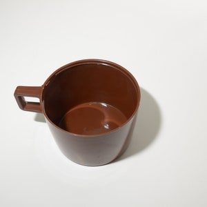 May include: A brown plastic cup with a handle. The cup is empty and sits on a white surface. The handle is rectangular and attached to the side of the cup.