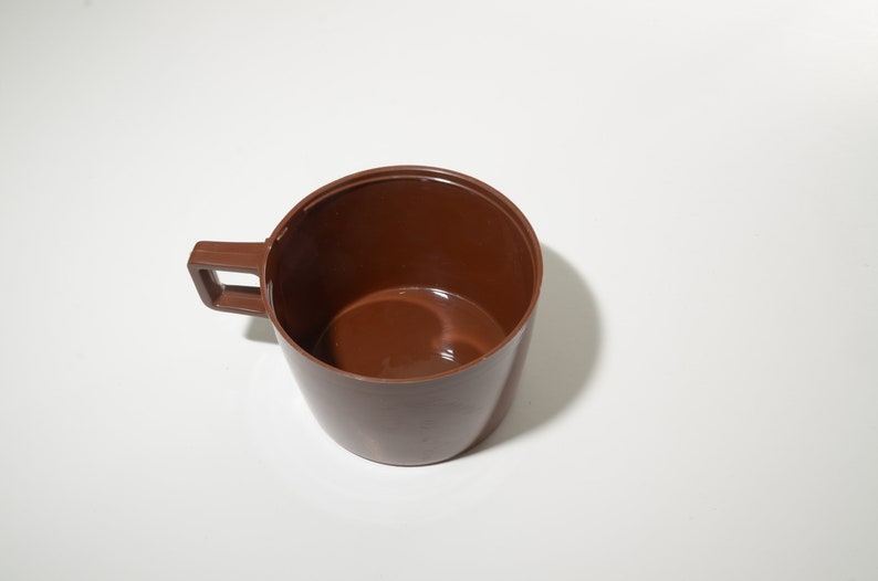 May include: A brown plastic cup with a handle. The cup is empty and sits on a white surface. The handle is rectangular and attached to the side of the cup.