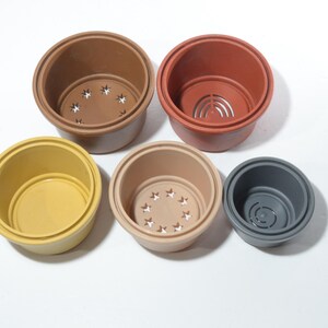 Puede incluir: Cinco macetas de plástico de diferentes tamaños y colores. Las macetas son marrones, amarillas, beige y grises. Cada maceta tiene un agujero de drenaje en la parte inferior.