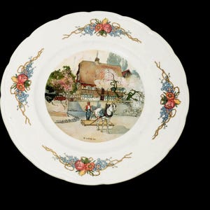 Puede incluir: Plato decorativo blanco con borde festoneado. El centro presenta una escena pastoral con un carro tirado por caballos, una cabaña y una persona. Acentos florales y de vid adornan el borde.