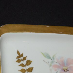 May include: A white rectangular dish with a gold rim and floral design. The dish features a pink flower with blue and green accents, and gold leaf detailing. The dish is likely porcelain.
