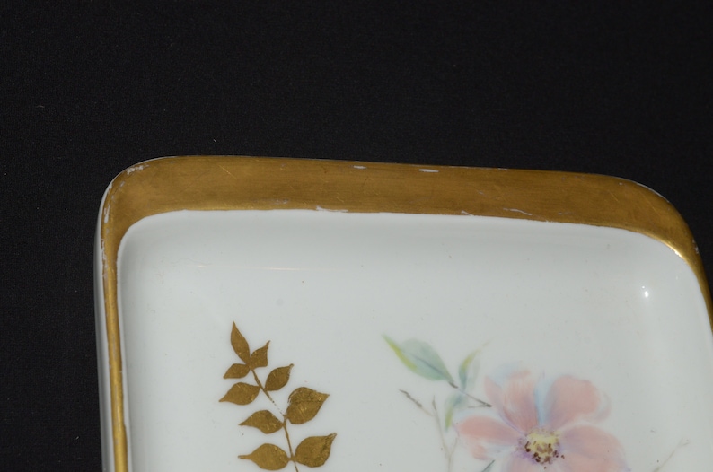 May include: A white rectangular dish with a gold rim and floral design. The dish features a pink flower with blue and green accents, and gold leaf detailing. The dish is likely porcelain.