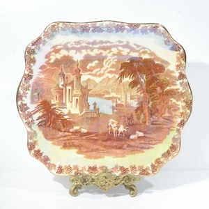 Peut inclure: Une assiette en porcelaine vintage à bord festonné présente une scène colorée d'un château, d'arbres et d'un lac. L'assiette est décorée d'un motif floral délicat et a un bord doré.